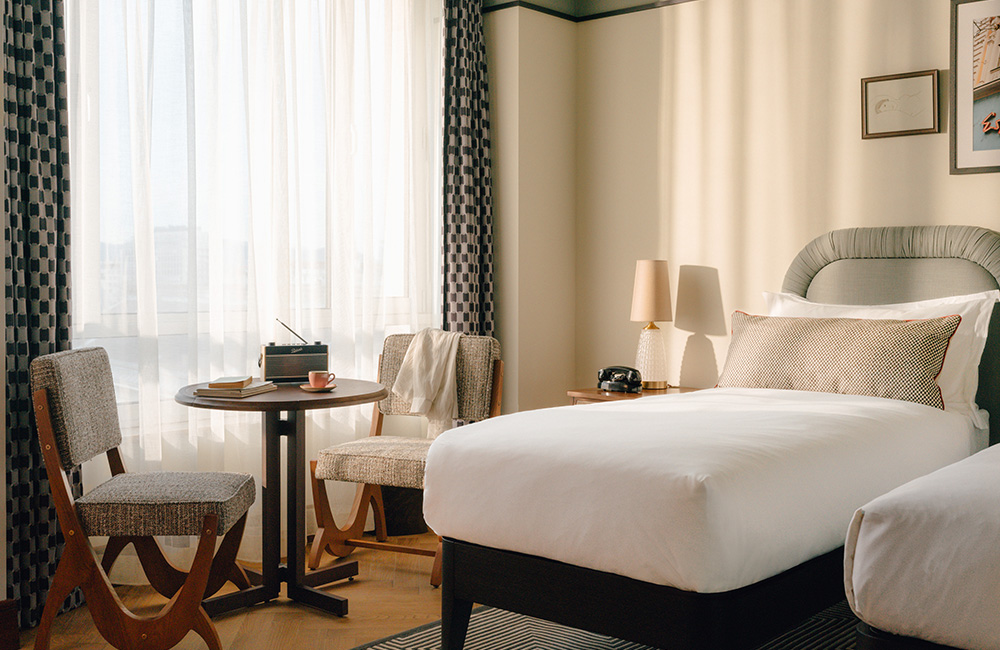 two single beds with chairs and table next to large window.